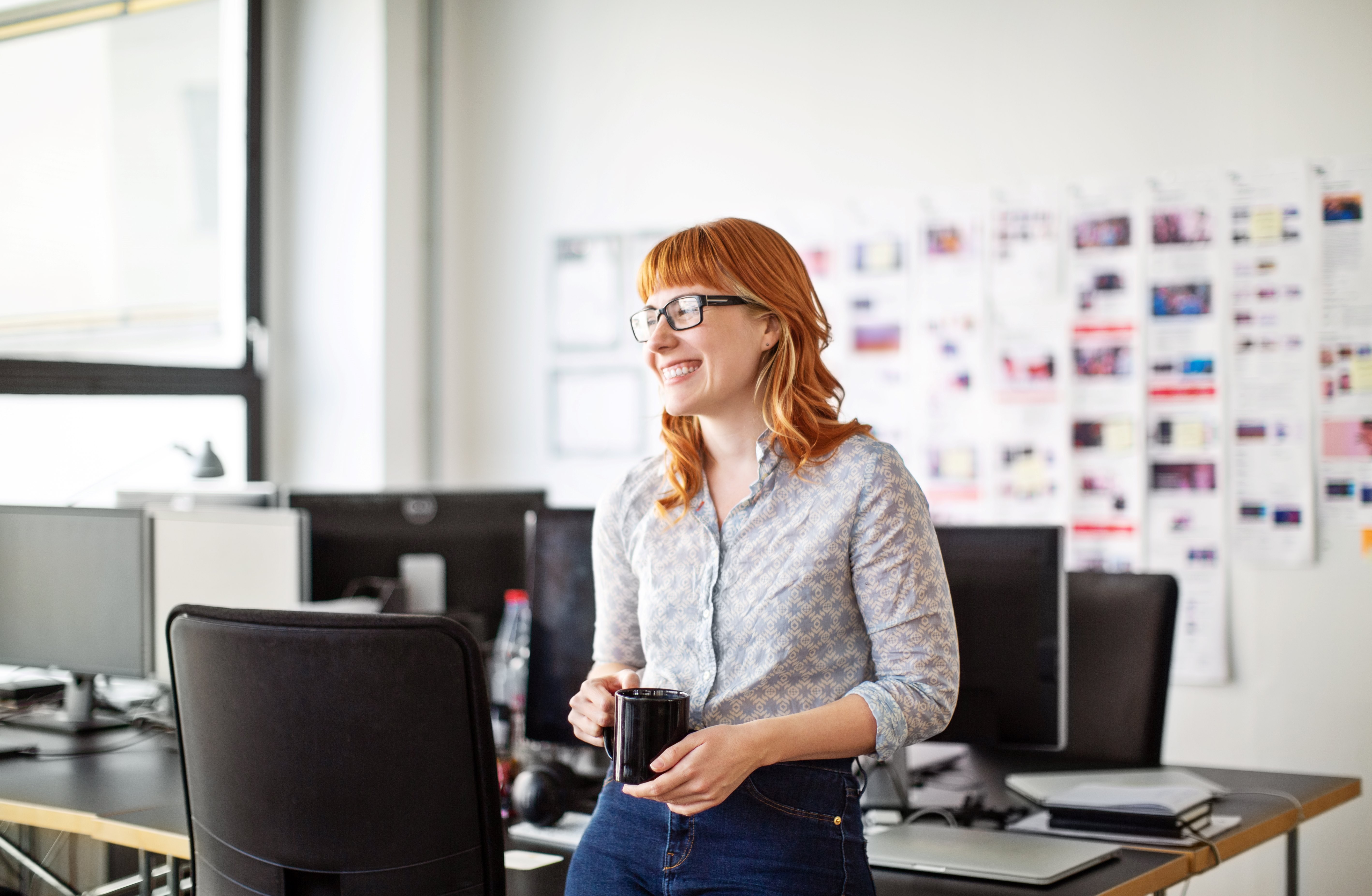 Woman with glasses smiling in an office, holding a coffee mug.