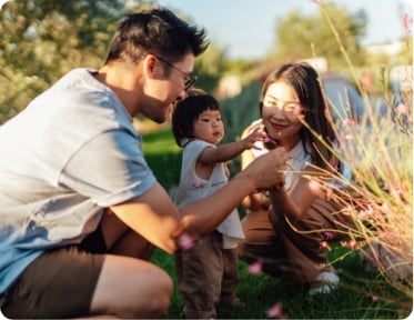 Two parents and a child in the park looking at a plant with flowers.