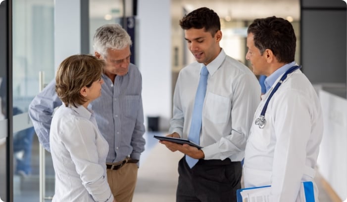 A doctor and man in a tie with a tablet speaking with an older couple.